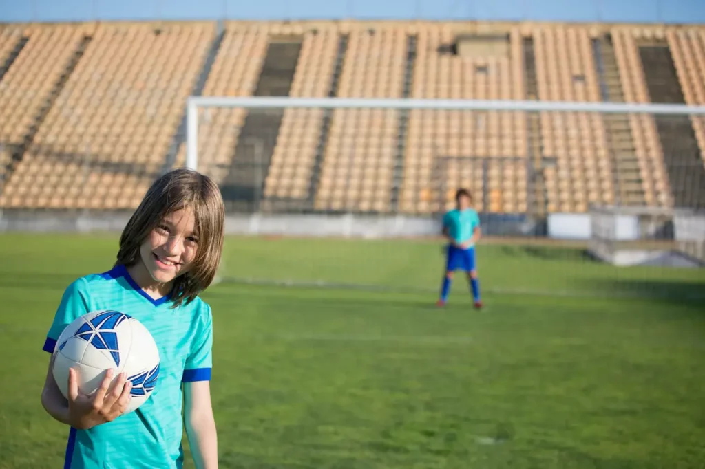 Smiling-girl-in-blue-jersey-proudly-holds-a-soccer-ball-with-an-expansive-stadium-and-a-distant-player-as-the-backdrop.-